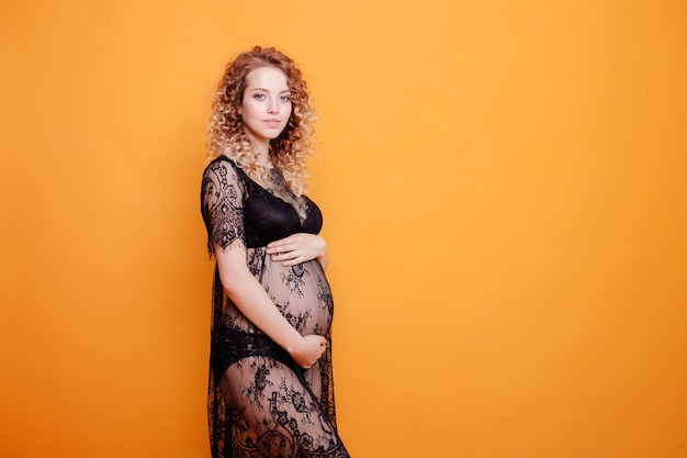 Pregnant young beautiful fashion blonde model woman in studio in black underwear on yellow orange background