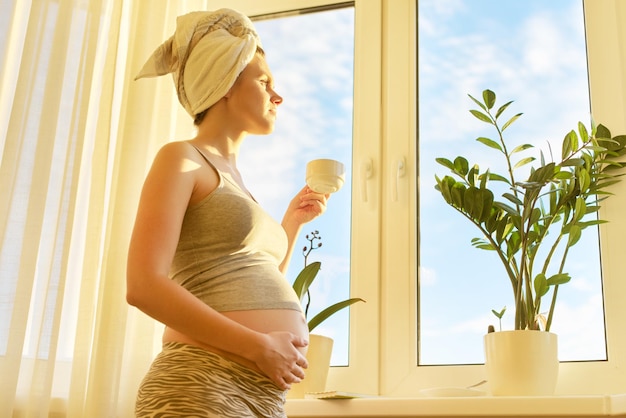 窓の外を見てお茶のカップと頭にバスタオルを持って妊娠中の若い女性