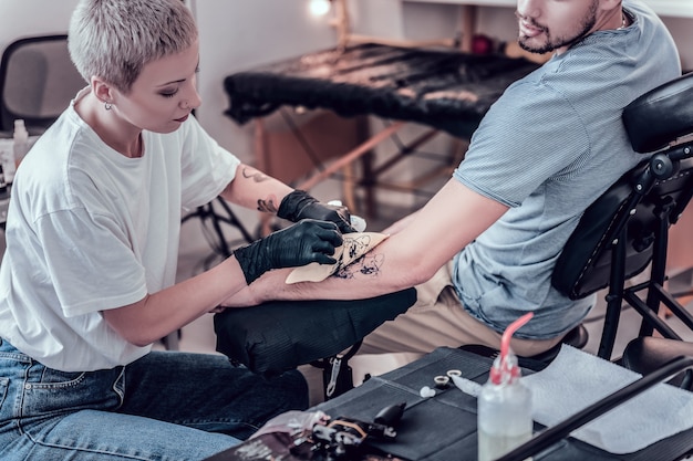 Предварительный эскиз. Внимательный татуировщик с короткой стрижкой аккуратно снимает бумажный эскиз с рисунком с поверхности кожи