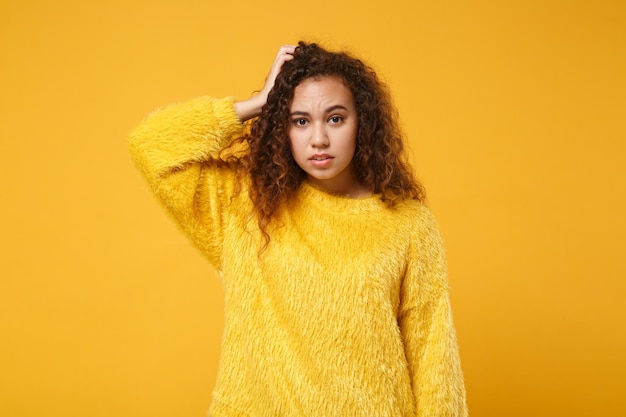 黄色オレンジ色の背景のスタジオの肖像画に分離された毛皮のセーターのポーズで夢中になっている若いアフリカ系アメリカ人の女の子。人々の誠実な感情のライフスタイルの概念。コピースペースをモックアップします。頭に手を置く。