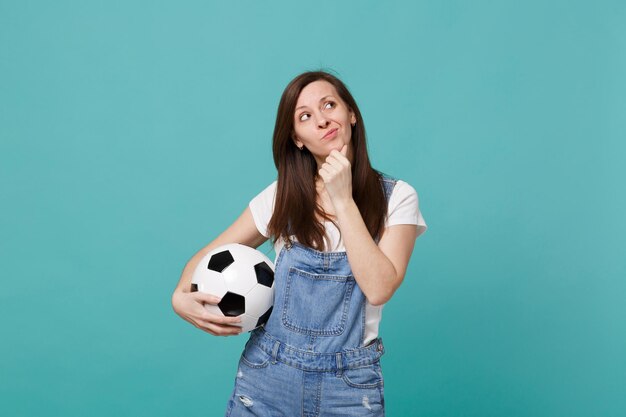 Preoccupied young woman football fan support favorite team with soccer ball looking up, put hand prop up on chin isolated on blue turquoise background. People emotions, sport family leisure concept.