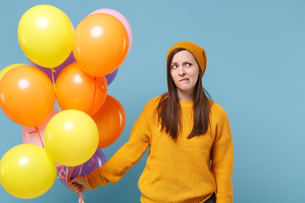 Preoccupied young woman girl in sweater hat posing isolated on\
blue background in studio. birthday holiday party people emotion\
concept. mock up copy space. celebrating hold colorful air balloon\
bitin