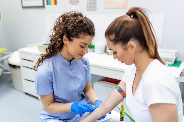 Preparazione per l'analisi del sangue con una bella giovane donna da parte di una dottoressa uniforme medica sul tavolo in una stanza bianca e luminosa l'infermiera trafigge la vena del braccio del paziente con un tubo vuoto dell'ago