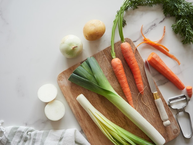 Preparation of healthy vegetable foods Onion potato leek and carrot on kitchen countertop on wooden cutting board next to knife and peeler Top view marble background