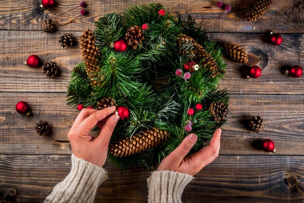 Preparation for xmas holidays. Woman decorating christmas green wreath with pine cones and red winter berries and christmas tree balls