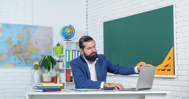 온라인 시험을 준비합니다. 수업에 대학 강사입니다. 학교로 돌아가다. 비정규 교육. 노트북 작업을 하는 심각한 성숙한 교사. 잔인한 수염 난 남자는 컴퓨터와 교실에서 작동합니다.