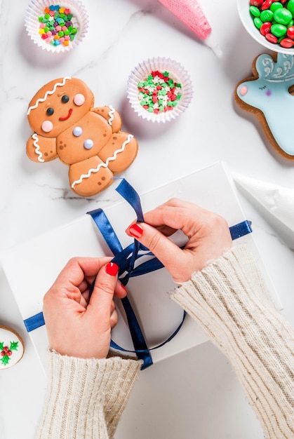 Preparing for christmas, decorating traditional gingerbread with multicolored sugar glaze, the girl folds the cookies in a white gift box, with ribbon bow, on a white marble table copyspace top view