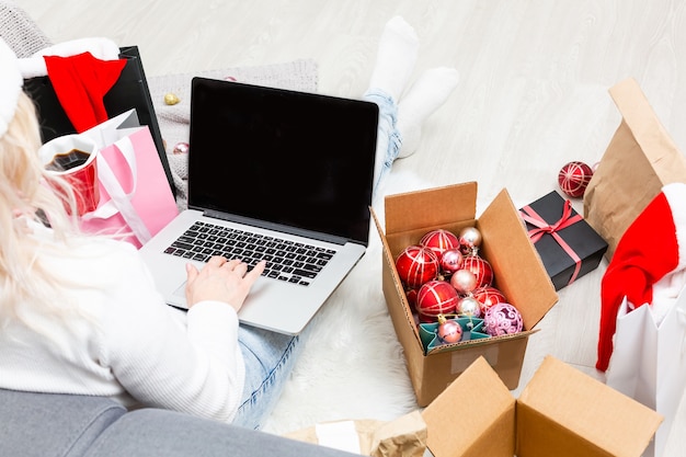 Preparing for Christmas party. Woman ordering presents and decorations on laptop, sitting among gifts boxes and packages, copy space