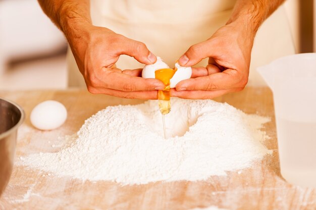 Preparing dough for pastry. Close-up of male hands cracking eggs upon flour