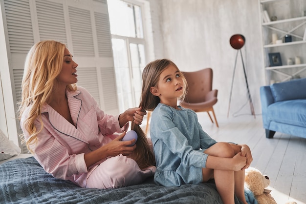 Preparing her to go. Beautiful young mother brushing her daughters hair while sitting on the bed at home