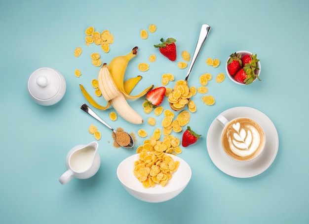 Preparing quick healthy breakfast in the morning: adding cereals, fruits into white bowl. Ingredients for making breakfast with granola, milk, freshly made cappuccino on blue background, top view set