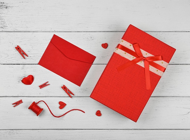 Подготовка подарков ко Дню святого Валентина, красная коробка, сердечки, шпагат, прищепки и записка в бумажном конверте на фоне белого деревянного стола, крупный план, плоский вид, вид сверху, прямо над