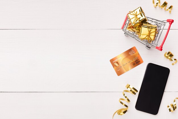 Preparing for winter holidays. Smartphone, credit card and trolley with gifts on white background, top view, copy space