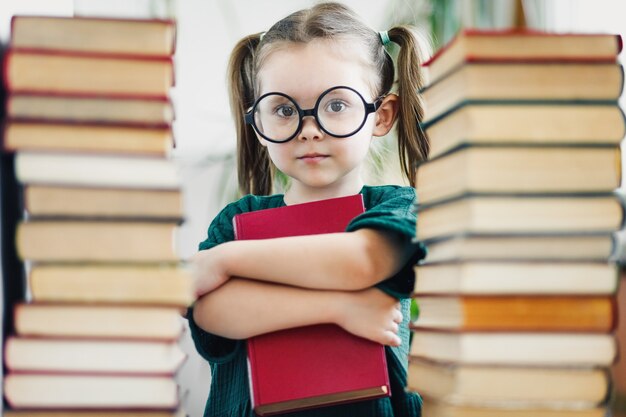 Preschool age girl in big round glasses holding red book among book piles.