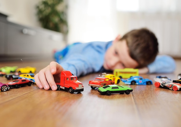 Preschool boy in a blue sweater is lying on the wooden floor playing with toy cars against the backg