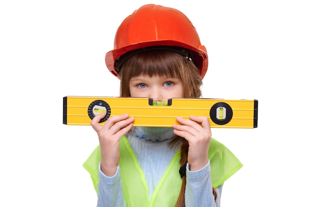 Девушка дошкольного возраста в строительном жилете и строительном шлеме держит уровень на белом изолированном фоне
