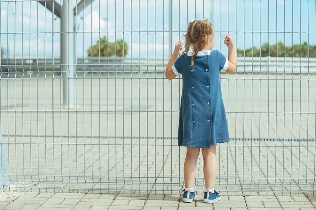 A preschool girl in a blue dress stands near a barrier from a high mesh fence and holds on to it