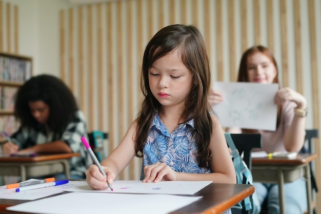 Disegno del bambino della ragazza in età prescolare con la matita di colore su carta bianca sul tavolo in aula con gli amici