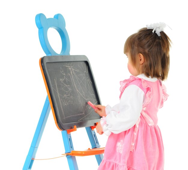 Preschool girl in pink dress draws with chalk on childrens board. Isolated on white wall