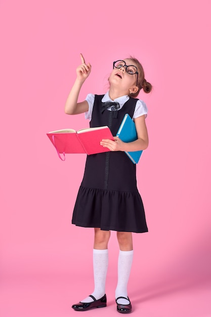 Дошкольная девочка в очках держит книгу, подняла руку и палец вверх на розовом пространстве