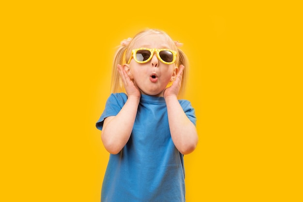 Preschool meisje in blauw T-shirt op gele achtergrond draagt een bril en maakt een verbaasd gezicht Studio portret