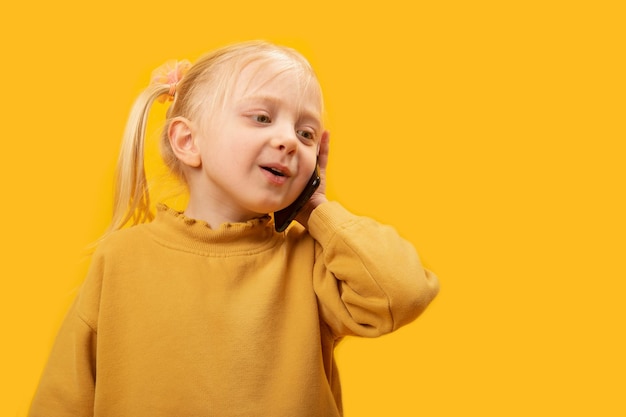 Preschool meisje in gele jas praten over telefoon Kinderen gebruik van smartphones Studio portret op gele achtergrond
