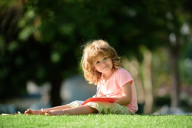 Preschool smiling pupil student boy writing to notebook in park on grass Home children learning Education childhood homework school concept