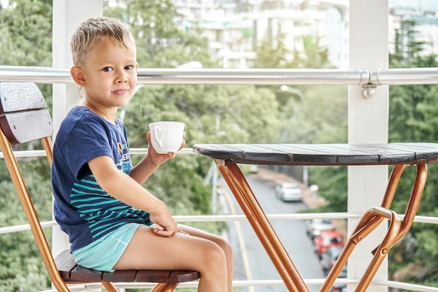 Preschooler boy sits drinking milk and enjoys life smiling sitting on chair on hotel balcony