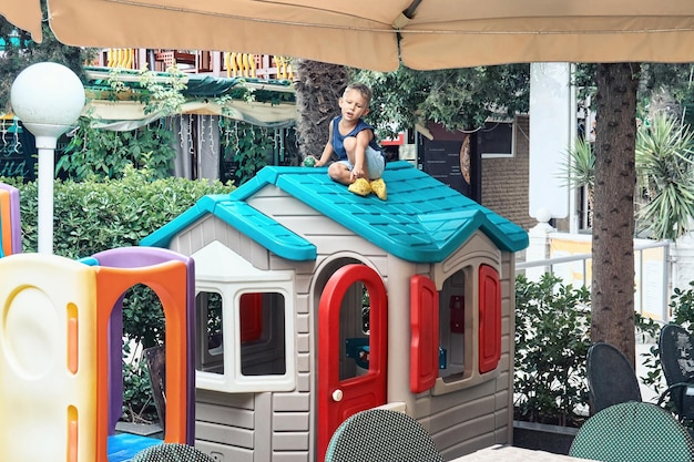 Preschooler boy in summer clothes sits on bright roof of toy house in restaurant play area