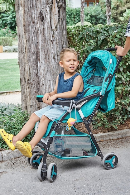 Preschooler frowns sitting in blue stroller of baby brother walking in park