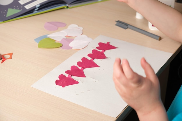 Preschooler girl in blue t-shirt makes applique sitting at the table, hearts cut out of colored bougue for card for Valentine's Day, children's creativity, atopic dermatitis on the hands of a child