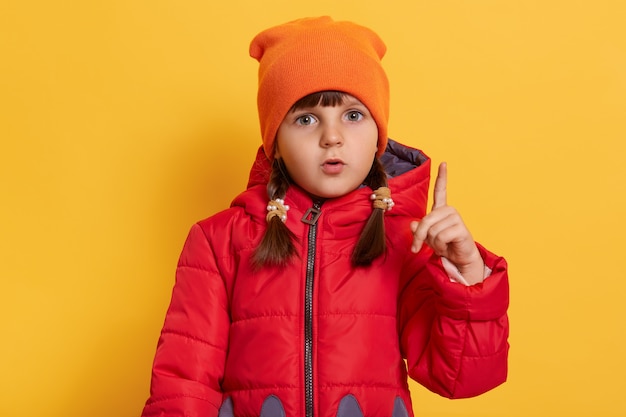 Preschooler girl shows index finger up isolated over yellow wall, has great idea, has thoughtful look, wearing red jacket and orange cap