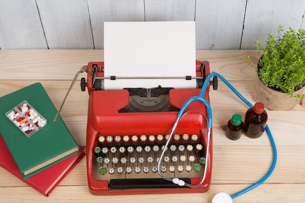 Prescription medicine or medical diagnosis - doctor workplace with blue stethoscope, pills, red typewriter with blank paper on wooden table