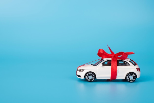 Present concept. Close up full length size photo of toy white car in with red ribbon isolated on blue background with copyspace