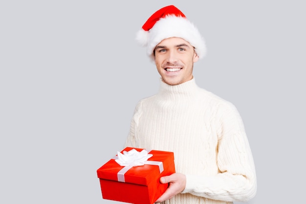 Present for you! Handsome young man in Santa hat holding a gift box and smiling while standing against grey background