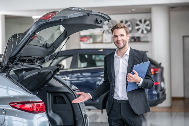 Presentation. Smiling man in dark suit and white shirt with folder pointing at open trunk of car standing in car dealership