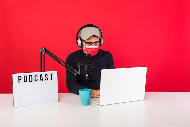 Presenter man wearing red cap and mask in podcast radio
recording studio, next to a computer, a microphone and a light box
with the word podcast, consulting the internet, on a red
background.