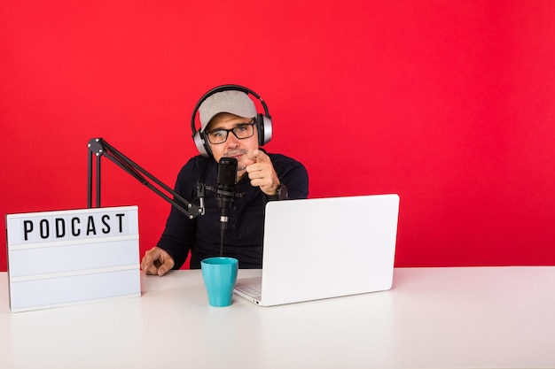 Presenter man with cap in podcast radio recording studio, next to a computer, a microphone and a light box with the word podcast, pointing to the audience, on red background.