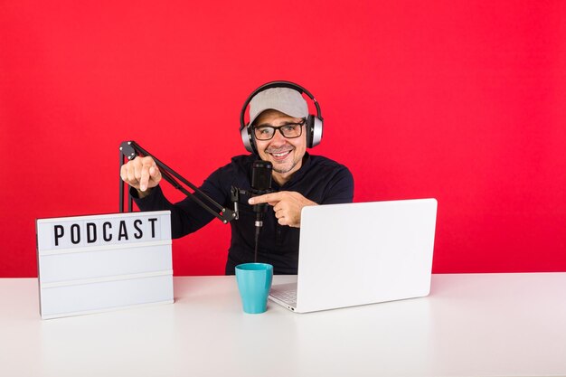 Presenter man with cap in podcast radio recording studio, next\
to a computer, a microphone and a light box with the word podcast,\
pointing to it, on a red background. podcasting, broadcast\
concept