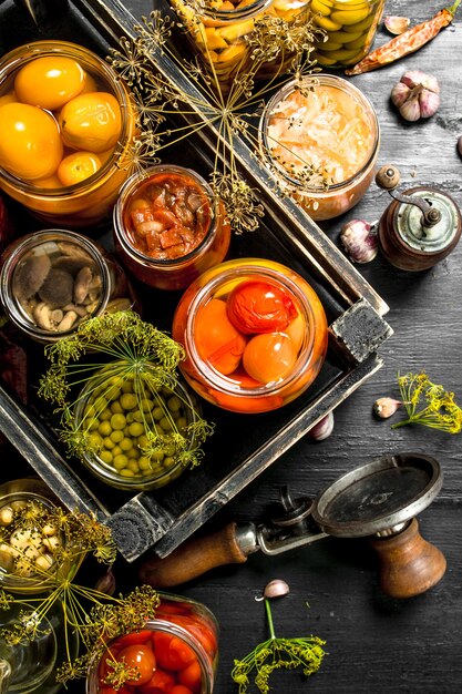 Preserved vegetables and mushrooms in a box with seamer on black chalkboard.