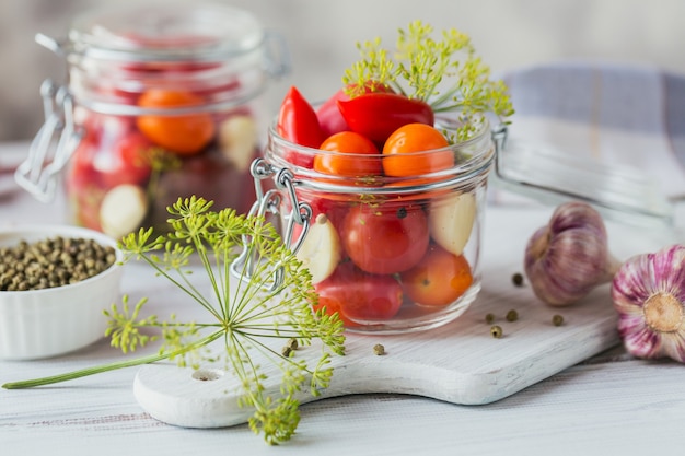 白い木製のテーブルにフレッシュトマトとピクルストマト、調味料、ニンニクを保存します。健康的な発酵食品。家庭用缶詰野菜。