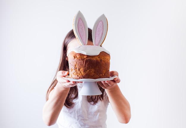 Ragazza preteen che tiene la torta di pasqua con glassa di zucchero e grandi orecchie da coniglio come parte della decorazione ragazza che copre il viso con pane pasquale su supporto bianco sfondo chiaro