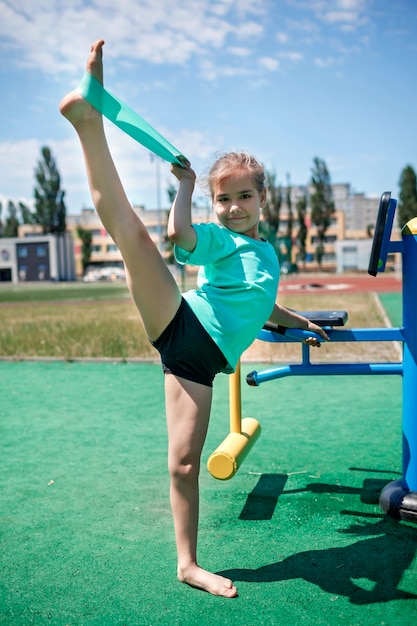 Preteen girl making exercises with fitness resistance band at public sportsground in city sport