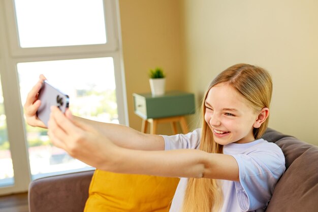 自宅の居心地の良いリビング ルームでスマートフォンで自撮り写真を作るプレティーンの女の子