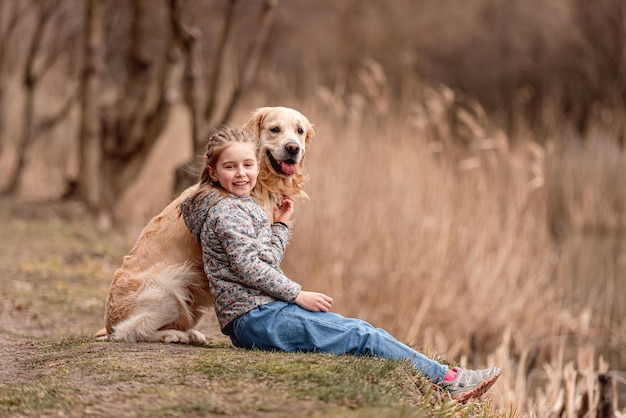 Preteen girl sitting with golden retriever dog on the nature and looking at the camera