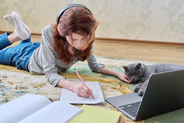 Preteen girl studying in headphones with laptop lying on floor with cat