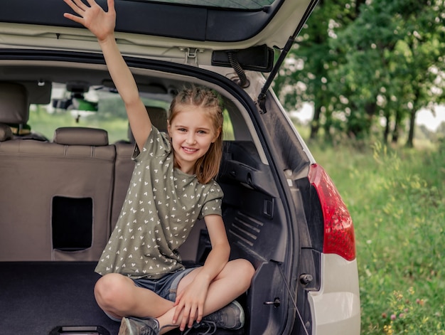 車のトランクに座って、手を上げて、カメラを見て笑っている美しい髪型のプレティーンの女の子。夏の旅行中に自然の中で車の中で幸せな子供子供