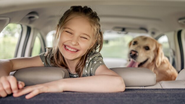 車の中に座って、カメラを見て、笑顔のゴールデンレトリバー犬とプレティーンの女の子。車内の純血種の犬のペットと子供