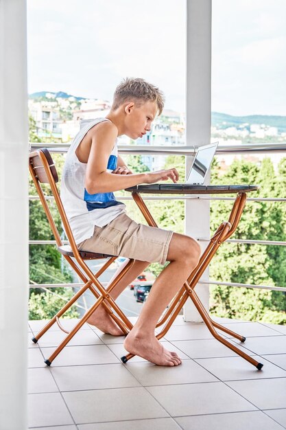 Preteen jongen zittend op het balkon van het hotel en surfen op het internet op grijze laptop