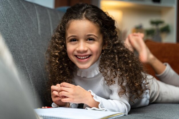 Preteen schoolgirl laughing to the camera while doing her homework at home Child study while writing something Education and distance learning for kids concept